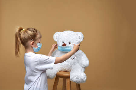 A little girl in a medical mask stands and puts a medical mask on her toy teddy bear.の写真素材