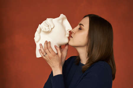 red-haired woman holds a piece of a plaster head in her hands and kisses it on a red background. The concept of psychology, falling in love, mental problems, relationshipsの写真素材