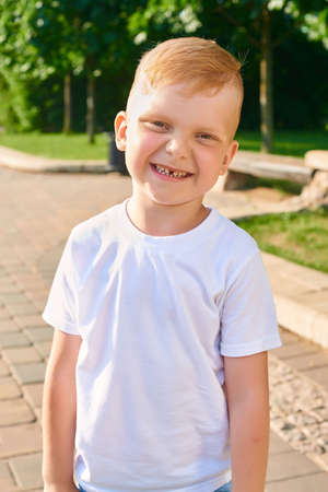 A small red-haired five-year-old boy stands in the park and smiles