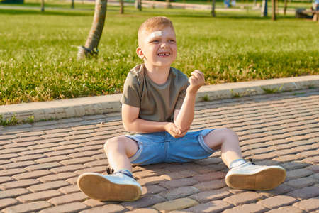 a small five-year-old red-haired boy fell, traumatized and felt pain. A child sits on a walkway and holds the elbowの写真素材