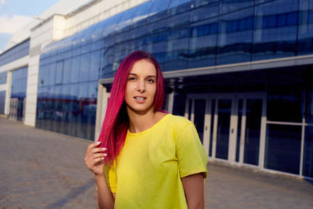 A pretty girl with colored dyed pink hair and a yellow T-shirt stands against the background of a glass buildingの写真素材
