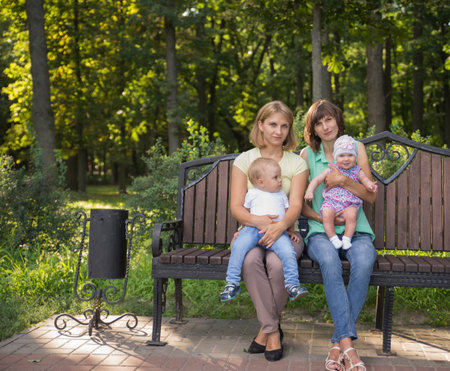 Two friends with children rest in the park in the summer. Sitting on a bench.の写真素材