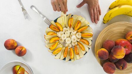 Apples, Bananas and Peaches Slices on a Plate. Woman Hands, Top View on White Background. Fruit salad, fruit platter, dessertの写真素材