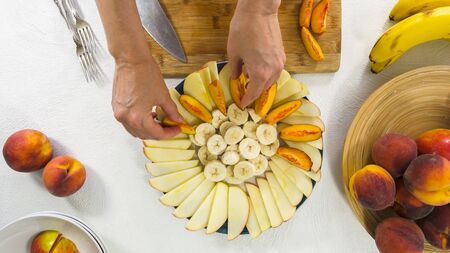 Apples, Bananas and Peaches Slices on a Plate. Woman Hands, Top View on White Background. Fruit salad, fruit platter, dessertの写真素材