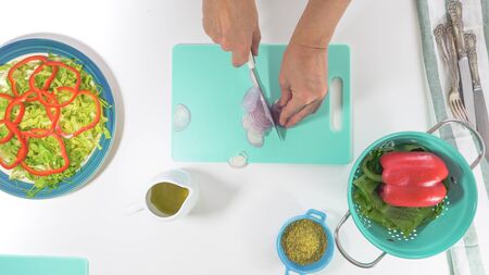 Onion slices on a cutting board. Vegetable salad on a plate, close up on white background. Fresh organic romaine lettuce, red pepper, onion, some seasoning, and olive oil. Top view, woman hands.の写真素材