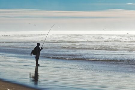 Saltwater fishing. Silhouette of man fishing on the beach. Stormy ocean, foggy dayの写真素材