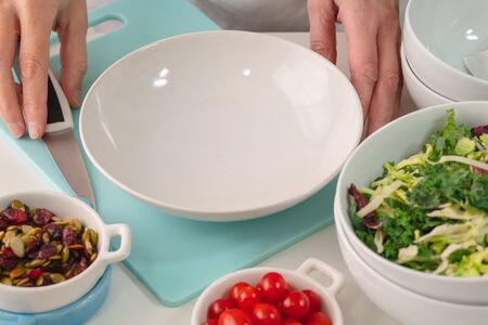 Woman preparing vegetable salad. Close up of fresh organic vegetables, cabbage mix, tomatoes, dried cranberries, and pumpkin seeds on a kitchen table on white background.の写真素材