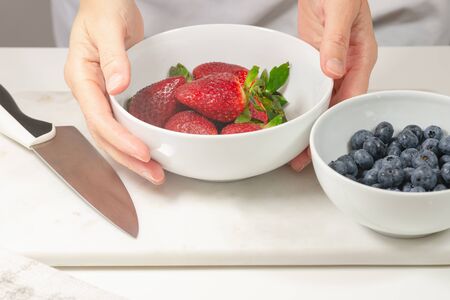 Strawberries and blueberries. Close up of fresh organic berries in a bowl, woman hands, white background. Woman preparing dessertの写真素材
