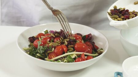 Preparation of vegetable salad. Woman puts pumpkin seeds and dried cranberries into a bowl with mix salad leaves and tomatoes, close up on white backgroundの写真素材
