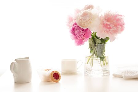 Pink peonies in glass vase, cup of tea, and cookies close up on white kitchen table in morning sunshine. Romantic breakfast, love, wedding, celebration conceptの写真素材