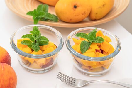 Chopped mangoes and peaches in a glass bowls with mint leaves close up on kitchen table. Fresh fruit salad, delicious dessertの写真素材
