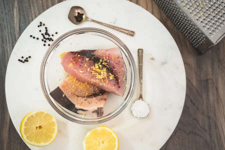 Raw swordfish marinated with salt, black pepper and lemon zest. Two pieces of fresh raw fish steaks in a bowl on marble cutting board with some ingredients, view from aboveの写真素材
