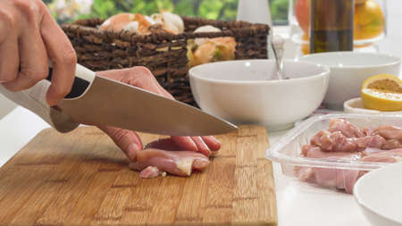 Woman chopping fresh raw chicken meat close up on a wooden cutting board on a kitchen tableの写真素材