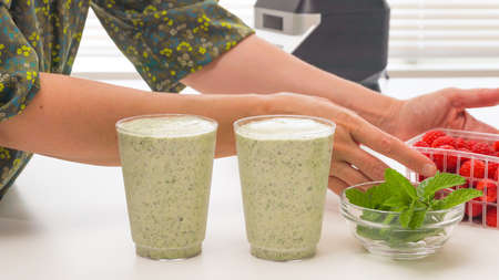 Celery banana smoothie with Chia seeds and honey in glasses, served with fresh raspberries and mint leaves, close up on kitchen table. Healthy breakfast, dessert, green detox smoothie recipeの写真素材