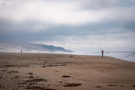 Foggy day on the beach. Silhouette of fisherman, flock of birds and dramatic cloudy sky on backgroundの写真素材