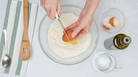 Ingredients for baking needs close up on light grey background. Woman's hands add eggs into a bowl...