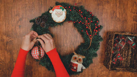 Woman decorating Christmas pine wreath with red winter berries, ornaments, and pine cones, close up view from above on wooden backgroundの写真素材