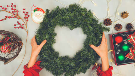 Woman decorating Christmas pine wreath with red winter berries, ornaments, and pine cones, close up view, flat lay, light grey backgroundの写真素材