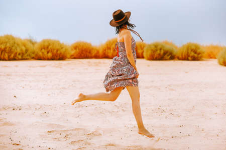 Happy woman in summer dress and hat running on sand beach in sunny day