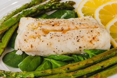 Cod fillet baked with garlic butter sauce served with fresh spinach and roasted asparagus on a white...