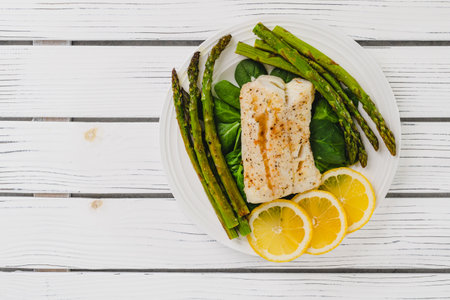 Cod fillet baked with garlic butter sauce served with fresh spinach and roasted asparagus on a white...