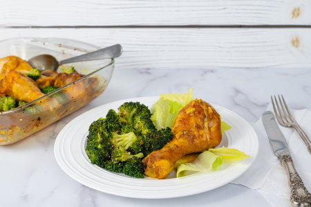 Serving dinner. Close-up view of a plate with an oven-baked chicken drumstick and broccoli served with fresh lettuce on a light marble backgroundの写真素材