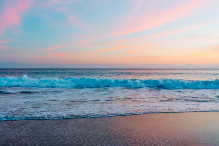 The sun sets over the tranquil beach, casting warm hues across the sky and reflecting its pink and golden glow on the gently lapping wavesの写真素材
