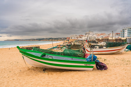 Fishing boats and equipment line the shore with a backdrop of a coastal town. Algarve, Portugal, January 3, 2024.のeditorial素材
