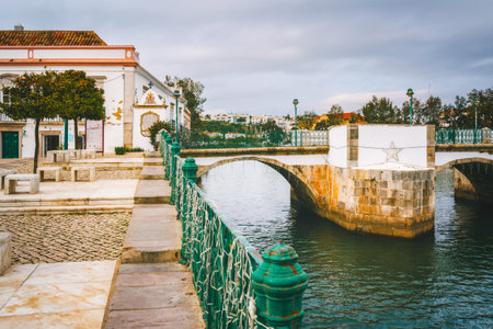 The Roman bridge in Tavira, Portugal, with white railings and stone arches, crossing over a calm...