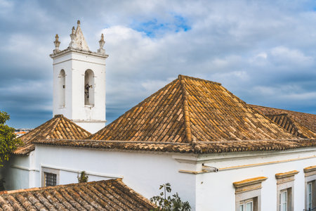A rooftop view in Tavira, Portugal, showing traditional terracotta roofs, a church tower, and a satellite dish under a cloudy sky.の写真素材