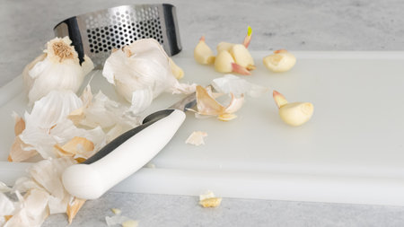 Peeled garlic, garlic press, and kitchen knife close-up on a white plastic cutting board on the kitchen tableの写真素材