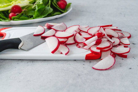 Sliced radishes on a white plastic cutting board, baby spinach, and lettuce on a light gray stone background, close-up viewの写真素材