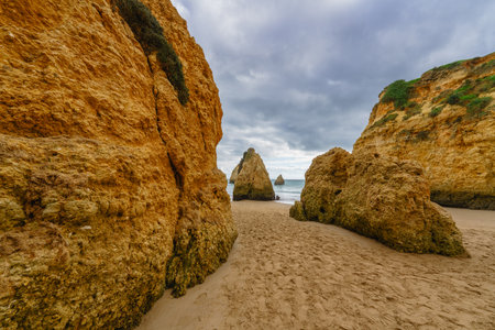 Rugged shore with scattered rocks, a sandy beach, and an overcast sky. Algarve coastline, Portugalの写真素材