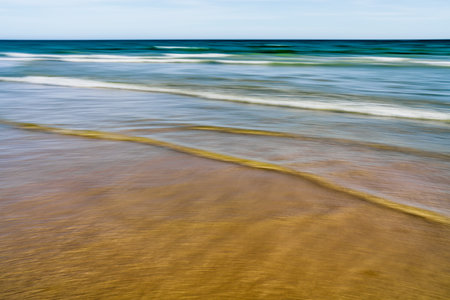 Blurred ocean waves merge into the sandy beach under a soft blue sky.の写真素材