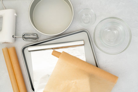 Flat lay composition featuring parchment paper, aluminum foil, a baking pan, mixer, and glass bowls arranged in a close-up view on a light grey background.の写真素材