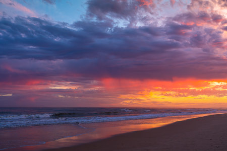 Waves roll in under a vibrant, dramatic sky painted with deep sunset hues.の写真素材