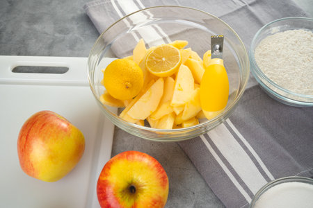 Baking pan with apple slices, lemon, eggs, sugar, and flour in bowls close-up on a kitchen table. Apple cake recipe, preparation processの写真素材
