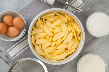 Baking pan with apple slices, eggs, sugar, and flour in bowls close-up on a kitchen table, flat lay. Apple cake recipe, preparation processの写真素材