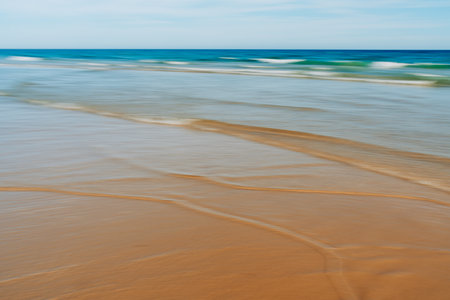 Gentle waves create soft patterns on a sandy beach with a blurred motion effect.の写真素材
