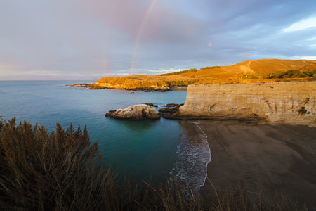 Rocks scattered along the beach are silhouetted against a vibrant sunset, with a stunning rainbow arching across the sky, California coastlineの写真素材
