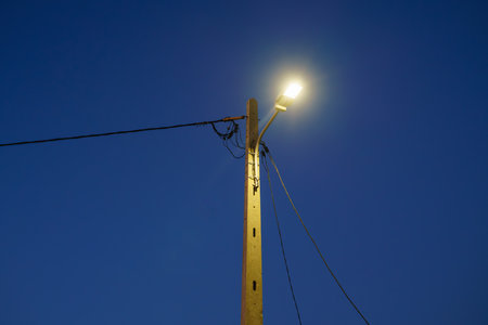 Illuminated streetlamp mounted on a wooden pole against a clear blue sky.の写真素材