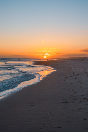 Vibrant sunset over a quiet beach with gentle waves and a lone figure near the water's edge.の写真素材