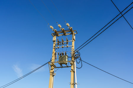 High-voltage power lines and transformer on a utility pole under a bright blue sky.