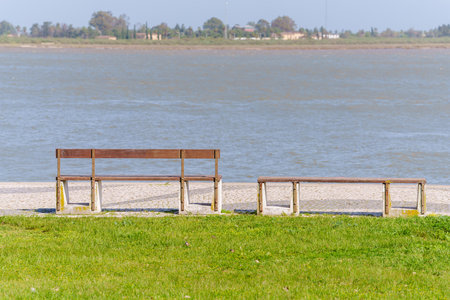 Two wooden benches, one with a backrest and one without, face a calm riverside view.の写真素材