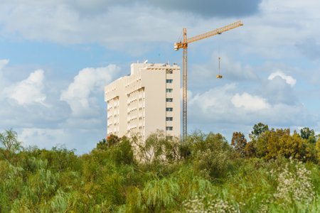 Tall beige building and construction crane rise above green trees under a cloudy sky.の写真素材