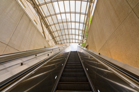 An upward view of an escalator ascending from an underground station with a modern glass roof and...