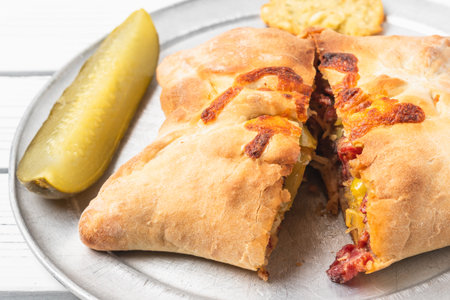 Golden-brown calzone stuffed with meats and vegetables served with a pickle spear on a silver tray.