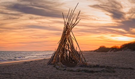 Tall wooden sticks form a teepee-like structure on a quiet beach during a vivid orange sunset.