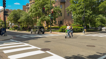 Washington D.C. USA - May 24, 2025. Two women on electric scooters cross a sunny street surrounded by historic architecture and greenery.のeditorial素材