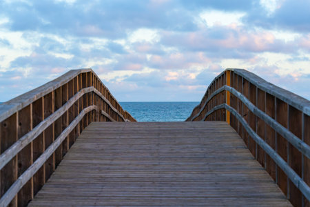 A wooden boardwalk curves gently toward the open ocean under a sky filled with soft clouds.